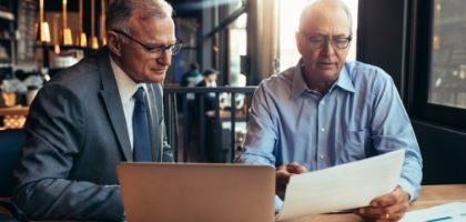 two men discussing paperwork. one holding a document and the other using a laptop