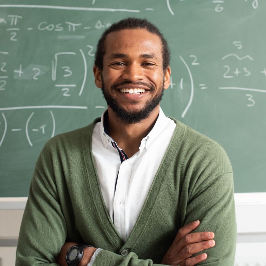 smiling male teacher standing in front of chalk board filled with equations 